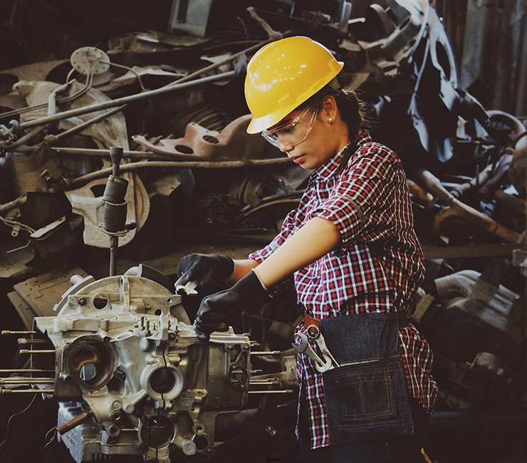 A female mechanic wearing a shiny yellow hard hat, a plaid shirt and wearing black gloves working on an engine.