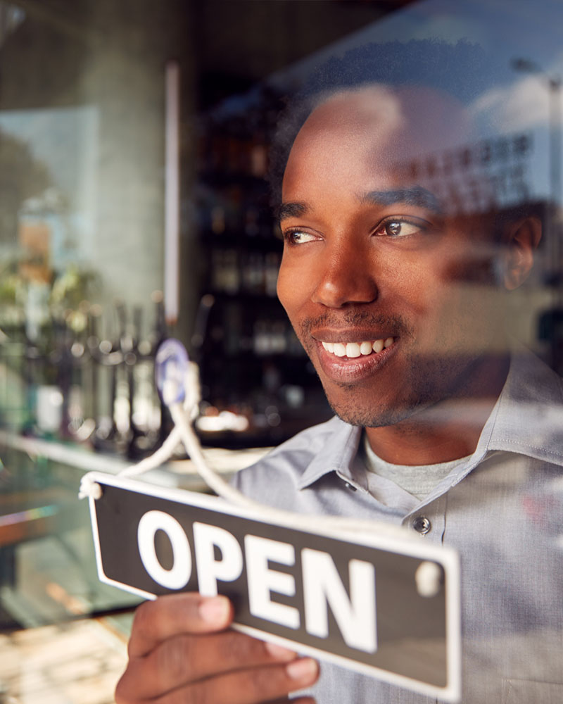 Picture of a Black business owner standing in front of his window flipping around a sign on the window to indicate he's open for business